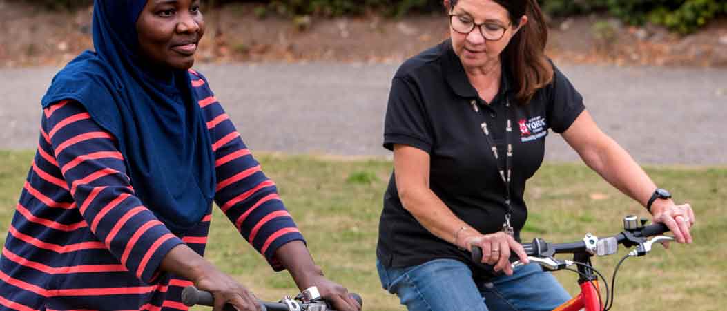 one lady is giving cycling lessons to another. There is grass and a pathway behind them.