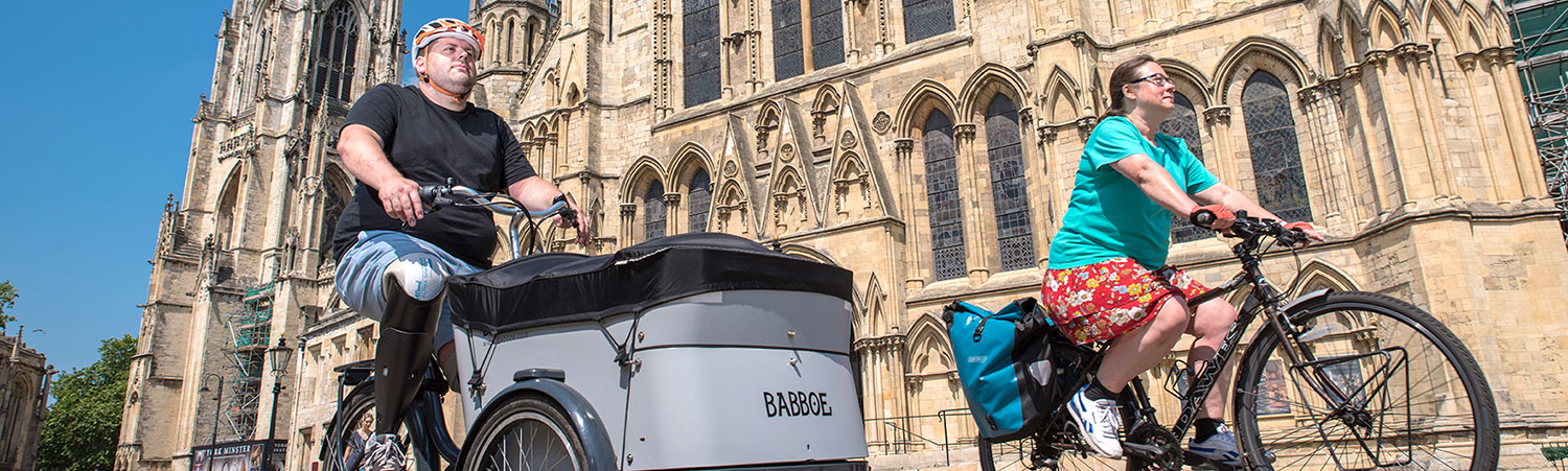 A disabled man and a women cycle past York Minster. The sun is shining and the sky is blue.